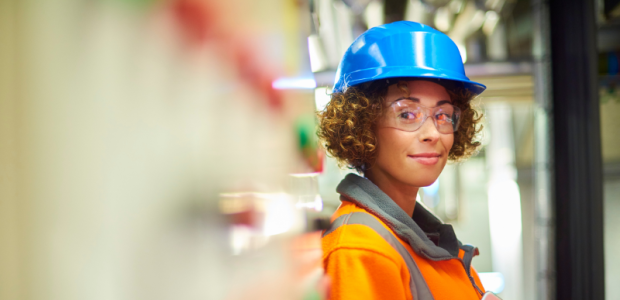 Trainerin während Schulung in der Produktionshalle Eine Trainerin mit orangener Weste und Schutzhelm gibt einem Schulungsteilnehmer Anweisungen in einer Produktionshalle. Im Hintergrund sind Maschinen und Produktionsanlagen zu sehen.